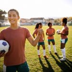 Porträt einer jungen schönen Frau, die Fußball spielt, im Hintergrund sieht man andere Spieler – © enigma_images - istockphoto.com - 1350461751